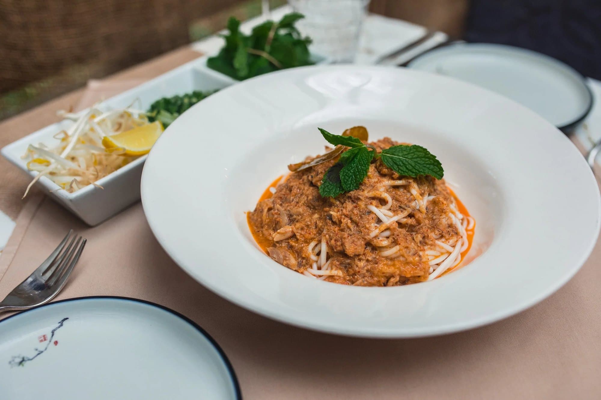 Wide white bowl of Bayon’s nyoam rice noodles topped with a thick red fish curry gravy and fresh mint, with bean sprouts and lemon on the side