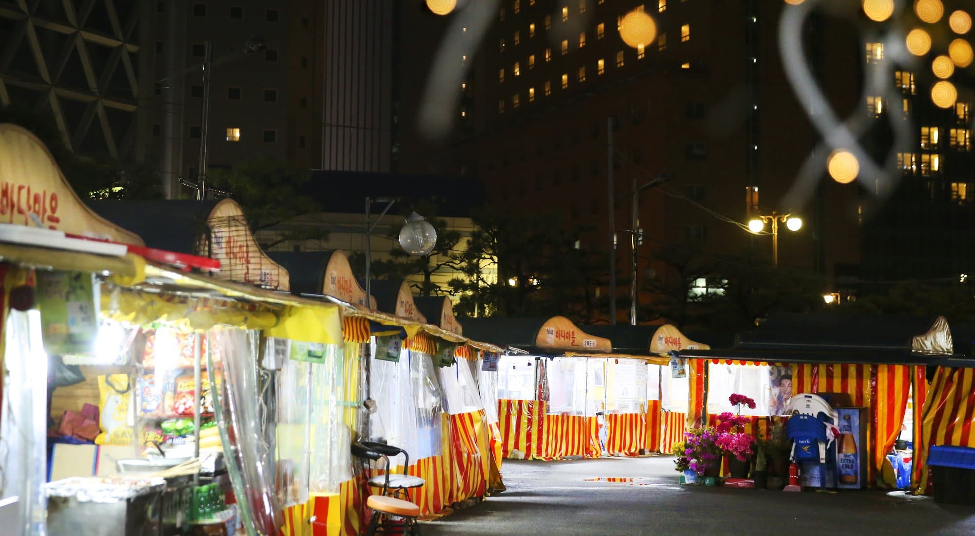 A night row of red-and-yellow pojangmacha tents with Hangul signs along a Busan street, city buildings behind them