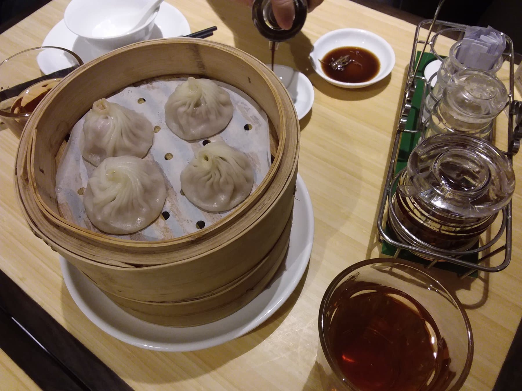 Bamboo steamer of four Crystal Jade xiao long bao on a wood table, with vinegar, ginger, and tea