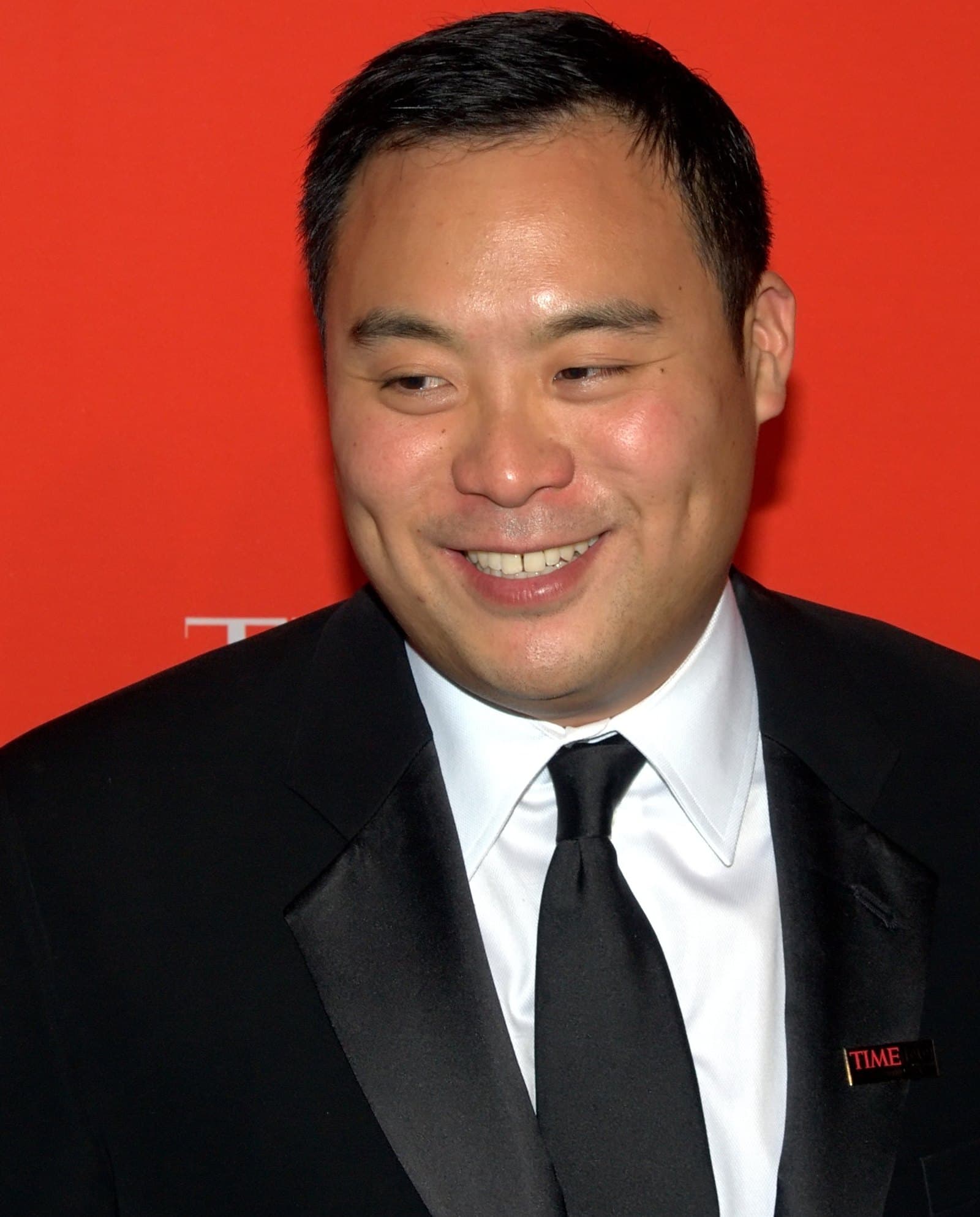 David Chang in a black tuxedo and white shirt, smiling against a red backdrop