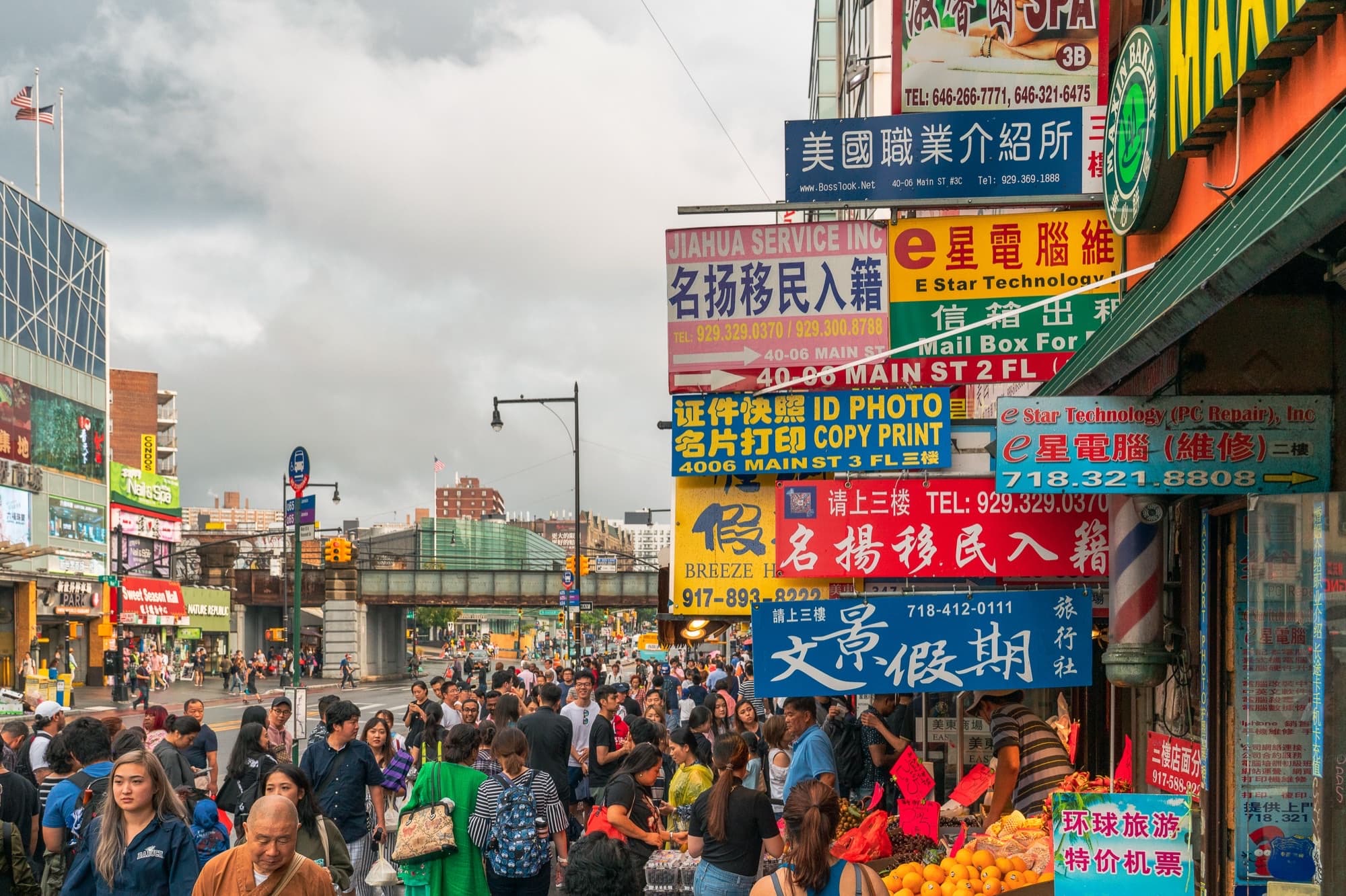 Pedestrians on a crowded Flushing Main Street sidewalk under layered Chinese and English commercial signs