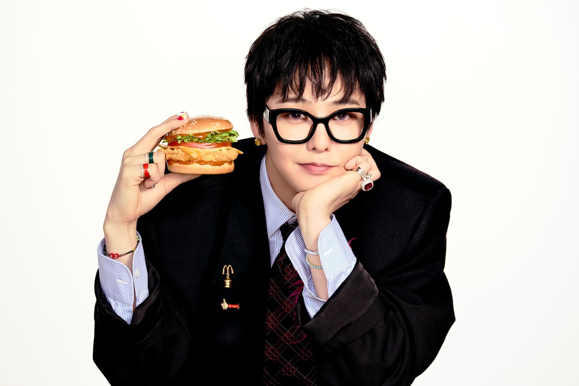 G-Dragon in a black blazer and glasses, chin on hand, holding a sesame-bun chicken sandwich from the McDonald’s gochujang butter campaign