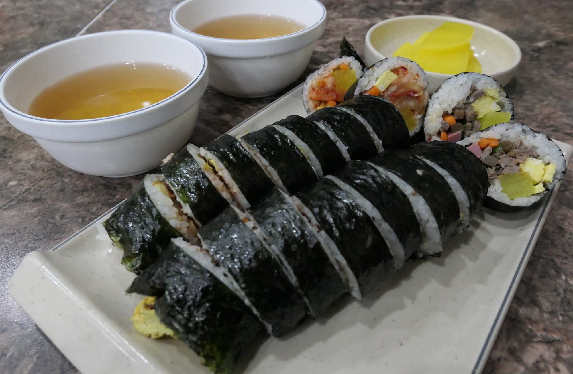 Two sliced gimbap rolls on a rectangular plate, with yellow pickled radish and small bowls of broth on a dark stone table