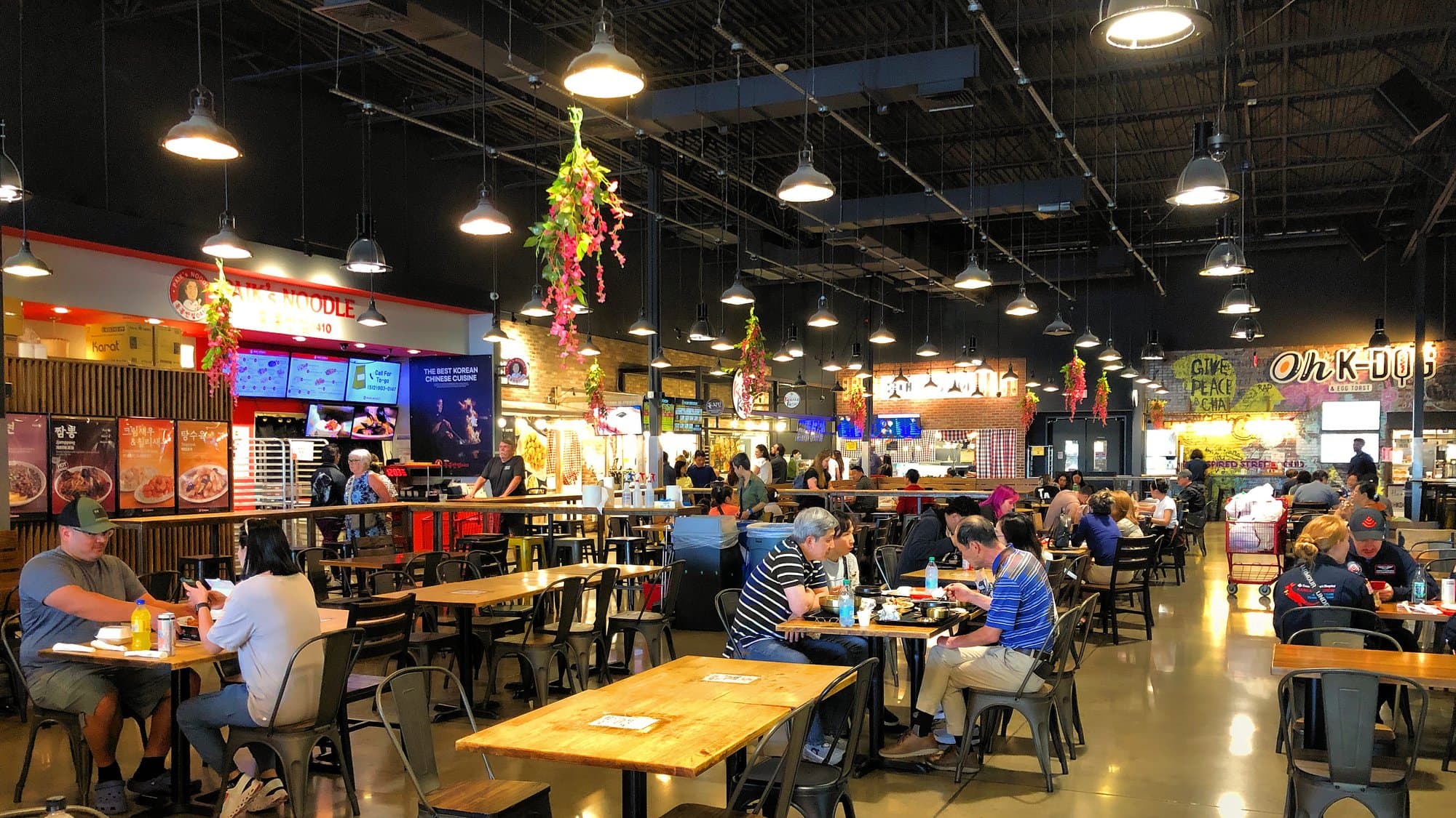 Diners at wooden tables inside H Mart’s Market Eatery in Austin, with noodle counters and hanging lights