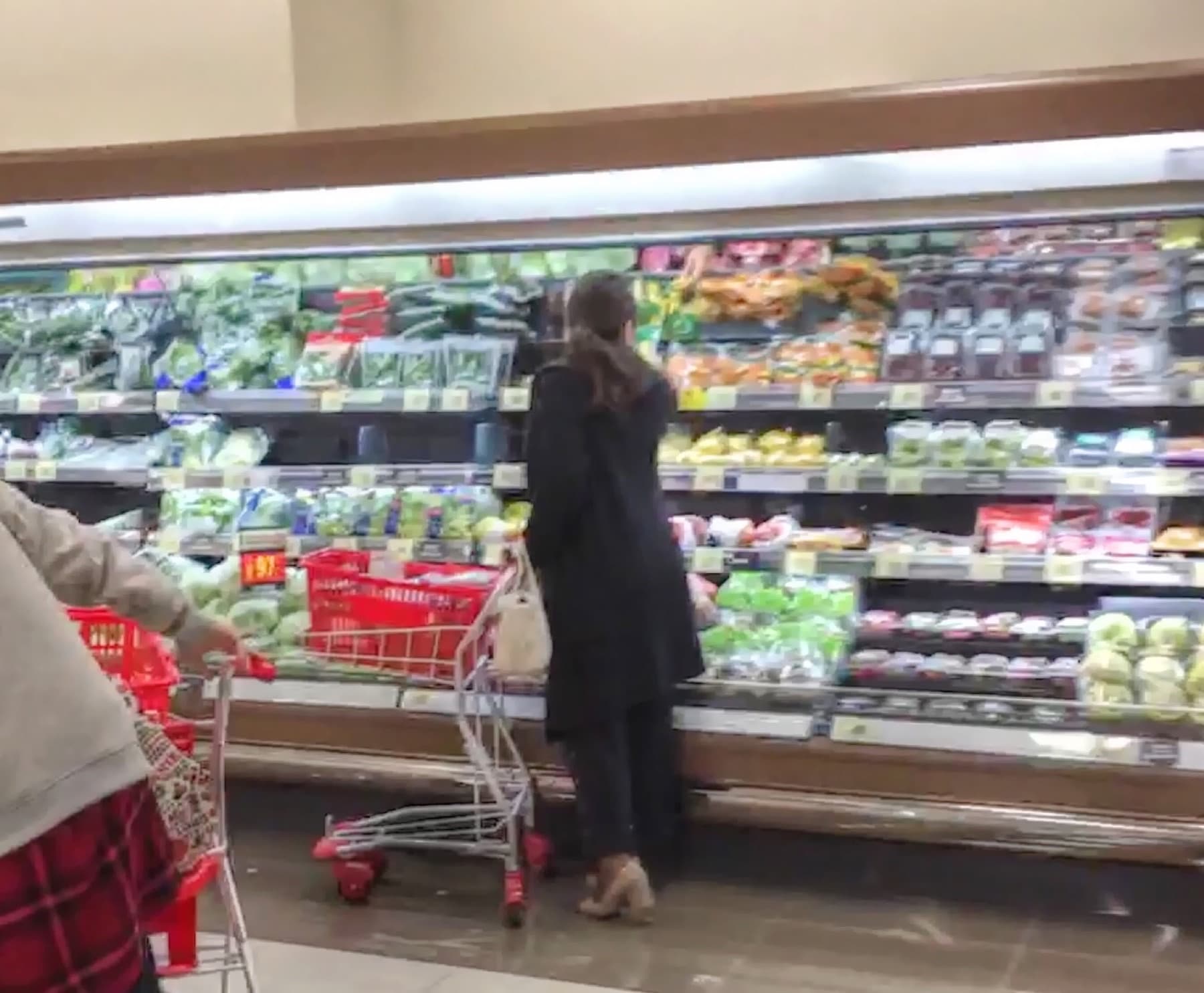 A shopper standing with a metal cart at refrigerated vegetable shelves in a Japanese supermarket