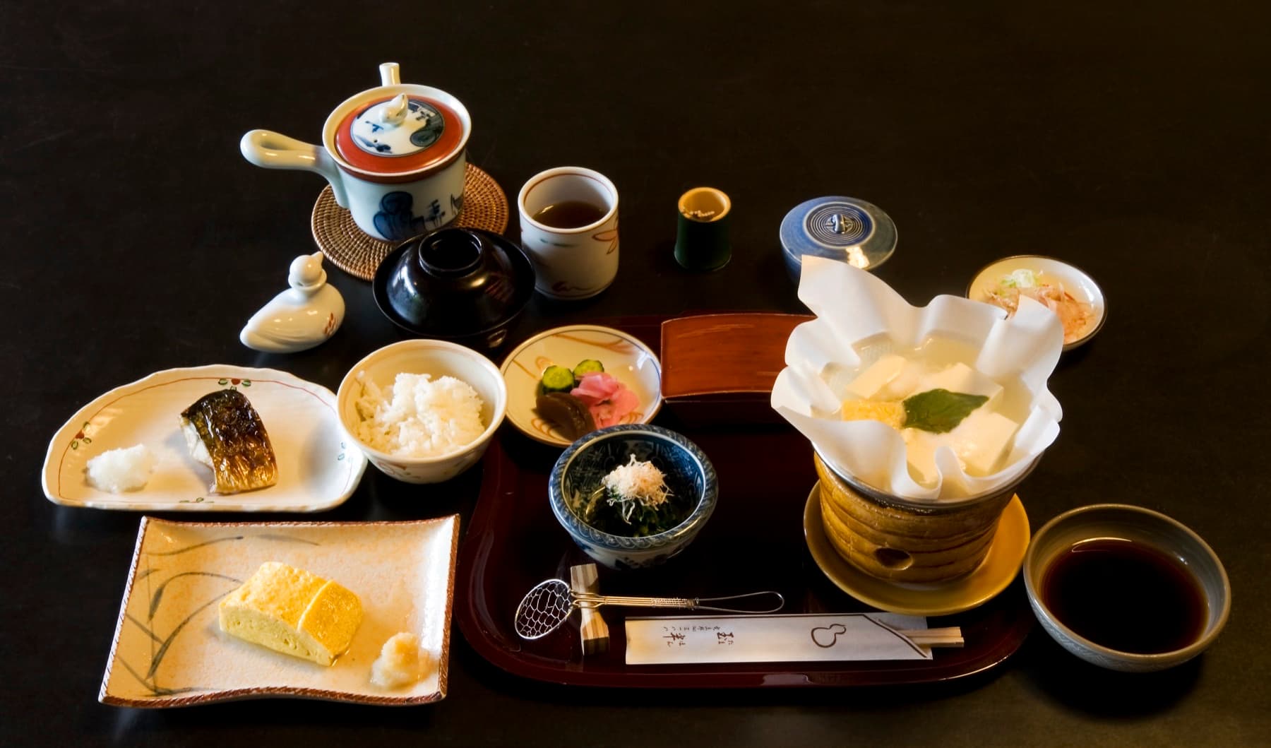 Japanese breakfast tray with grilled mackerel, rolled omelet, rice, miso soup, simmered tofu, pickles, and tea