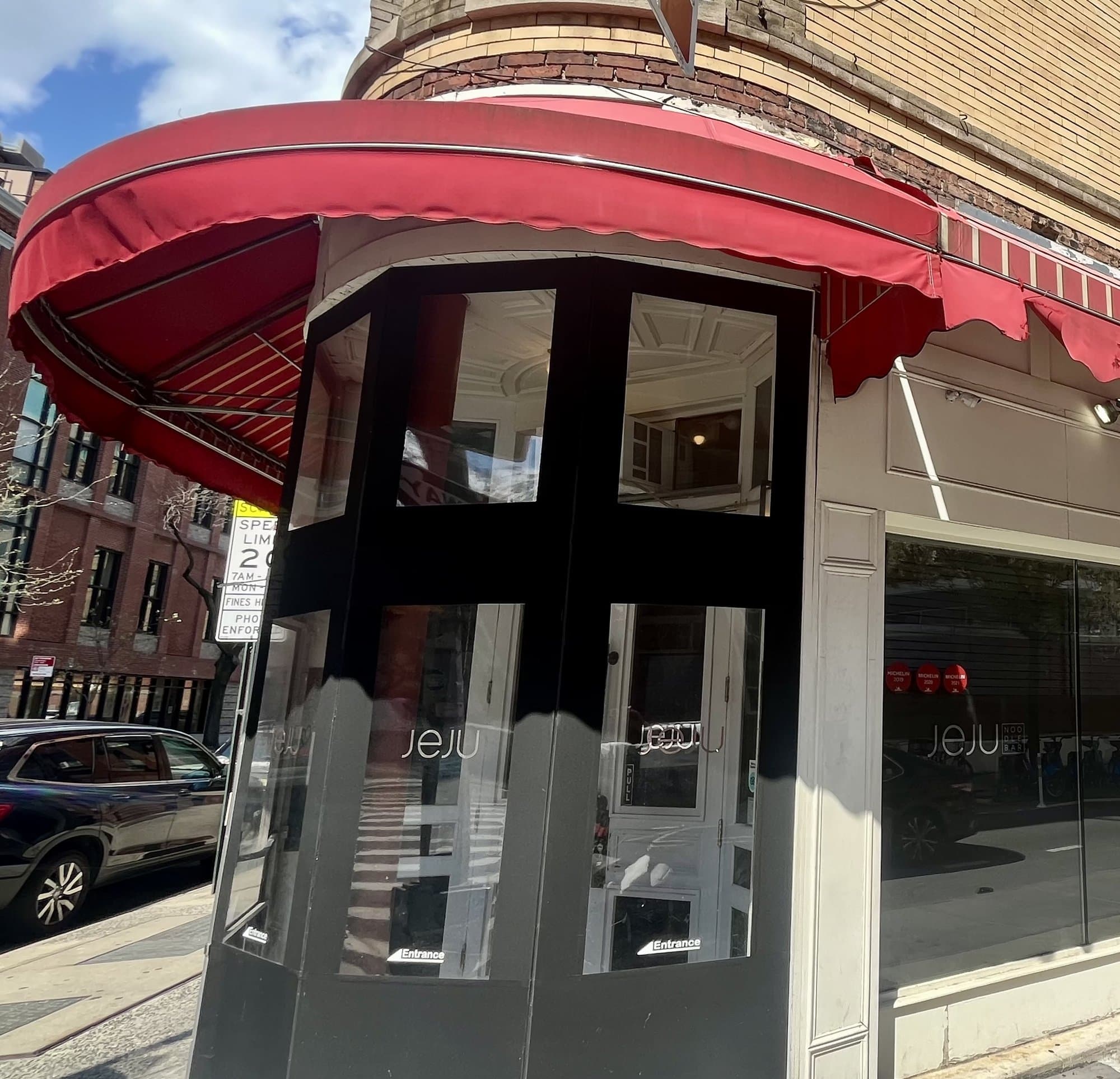 Corner door of Jeju Noodle Bar on Greenwich Street under a red awning, with the name on the glass