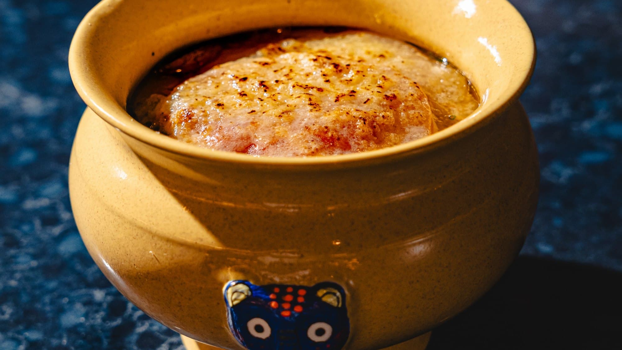 A yellow ceramic bowl of onion ramyun topped with a browned cap of cheese, with a small blue bear on the side, from Jeju Noodle Bar in Nolita
