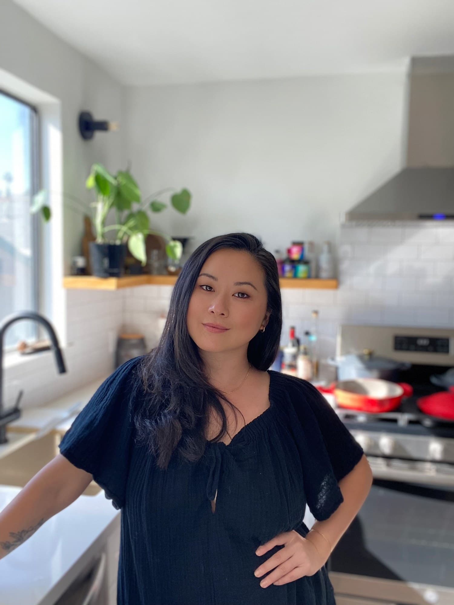 Jing Gao in a black dress standing in a bright kitchen, with a red Dutch oven on the stove behind her