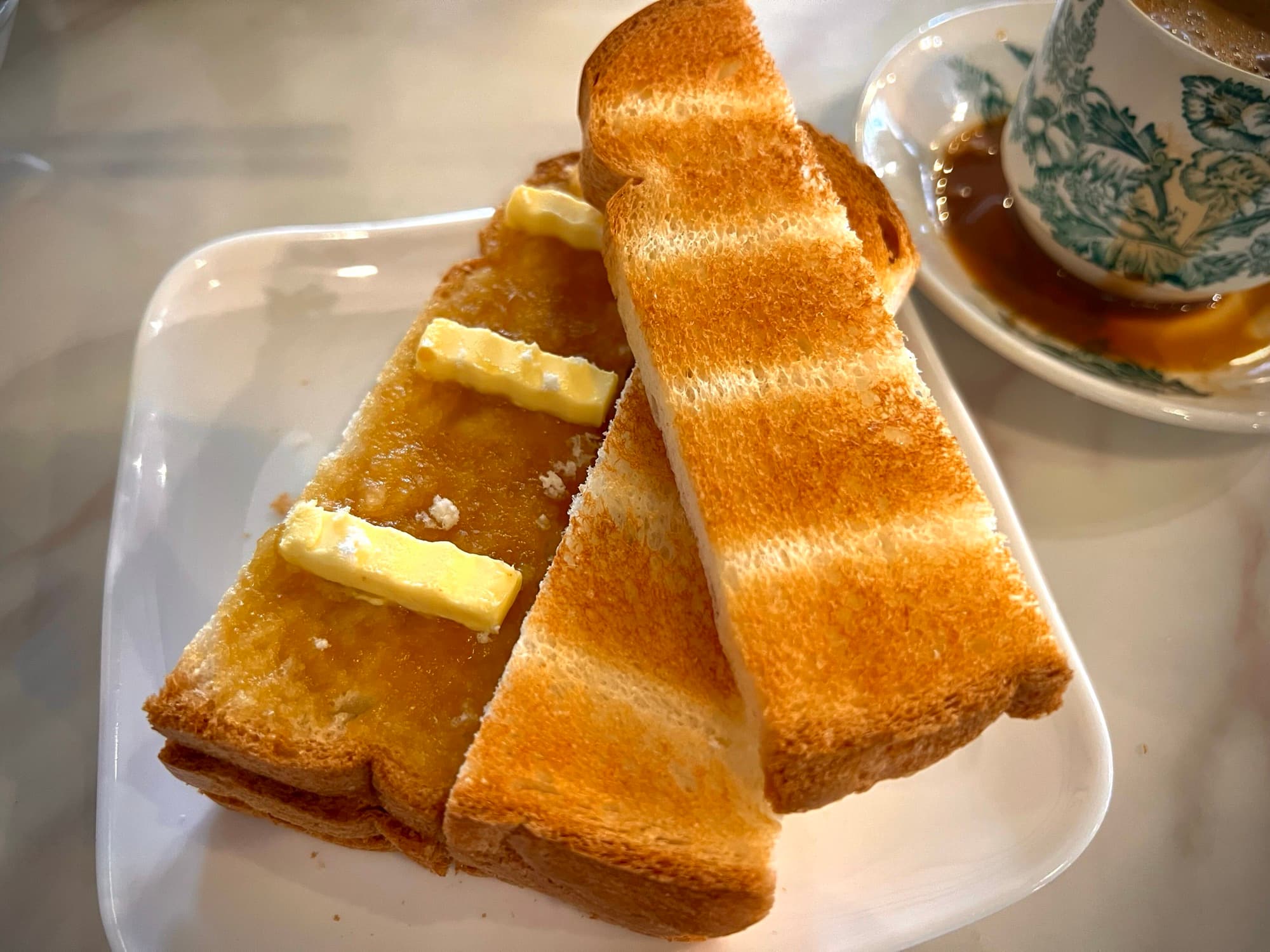 Thin grilled kaya toast topped with butter pats, beside a green floral cup of kopi on a marble table
