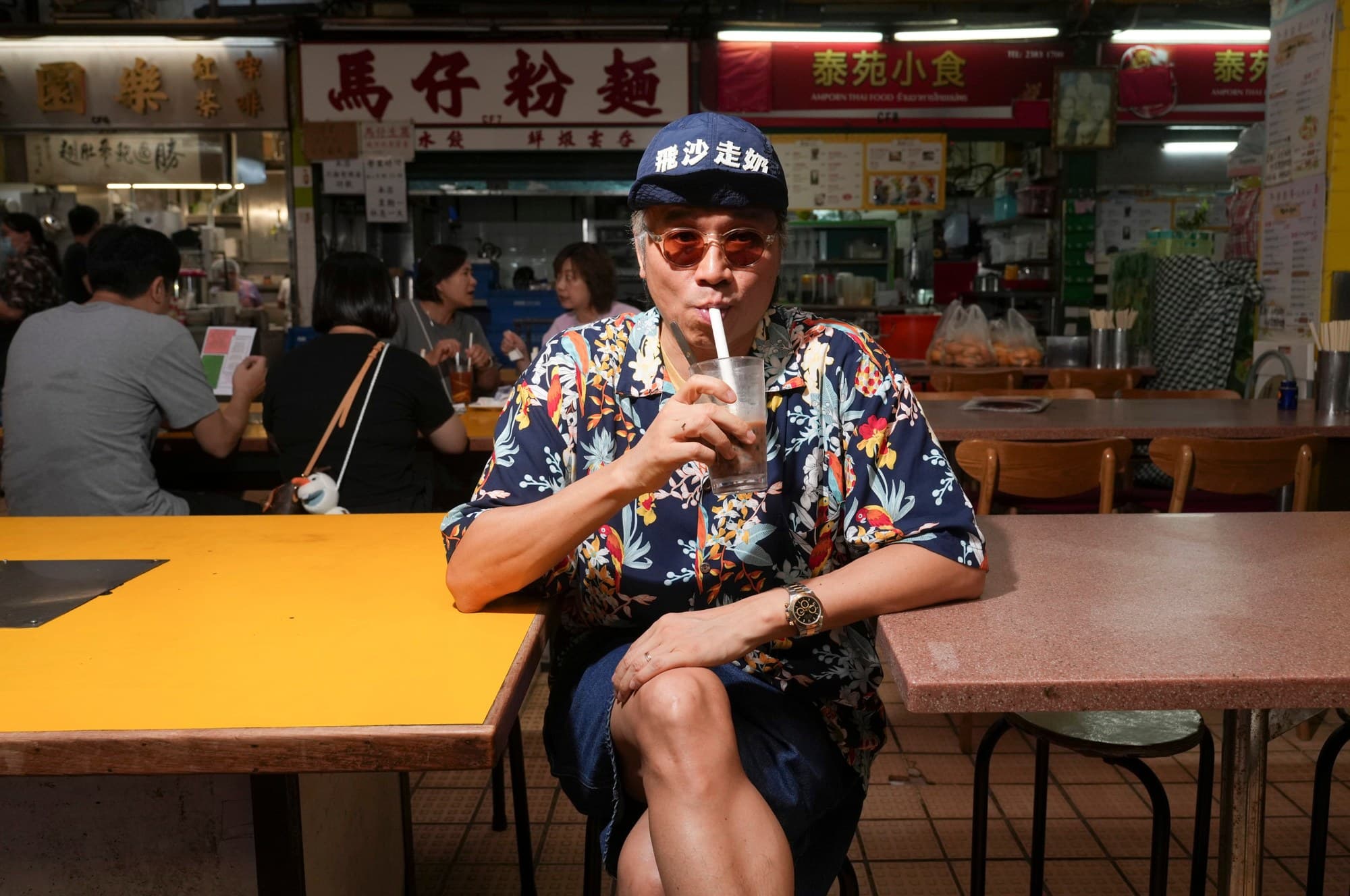 Kent Wong, in a blue cap and tinted glasses, drinking milk tea through a straw at a Hong Kong cooked-food centre