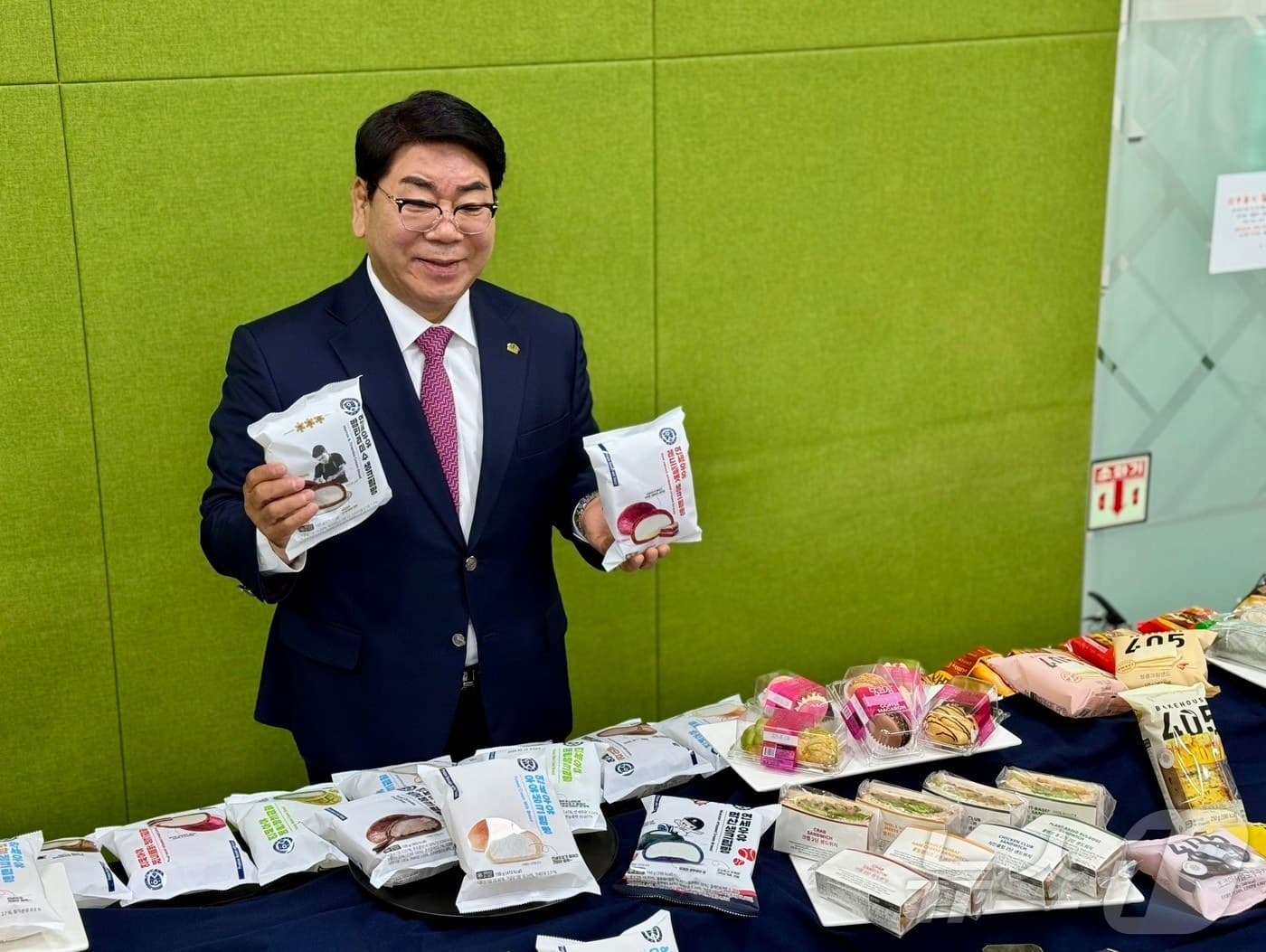 Foodcoa founder Kim Young-sik in a suit and glasses, holding two packaged cream breads behind a table of bakery products