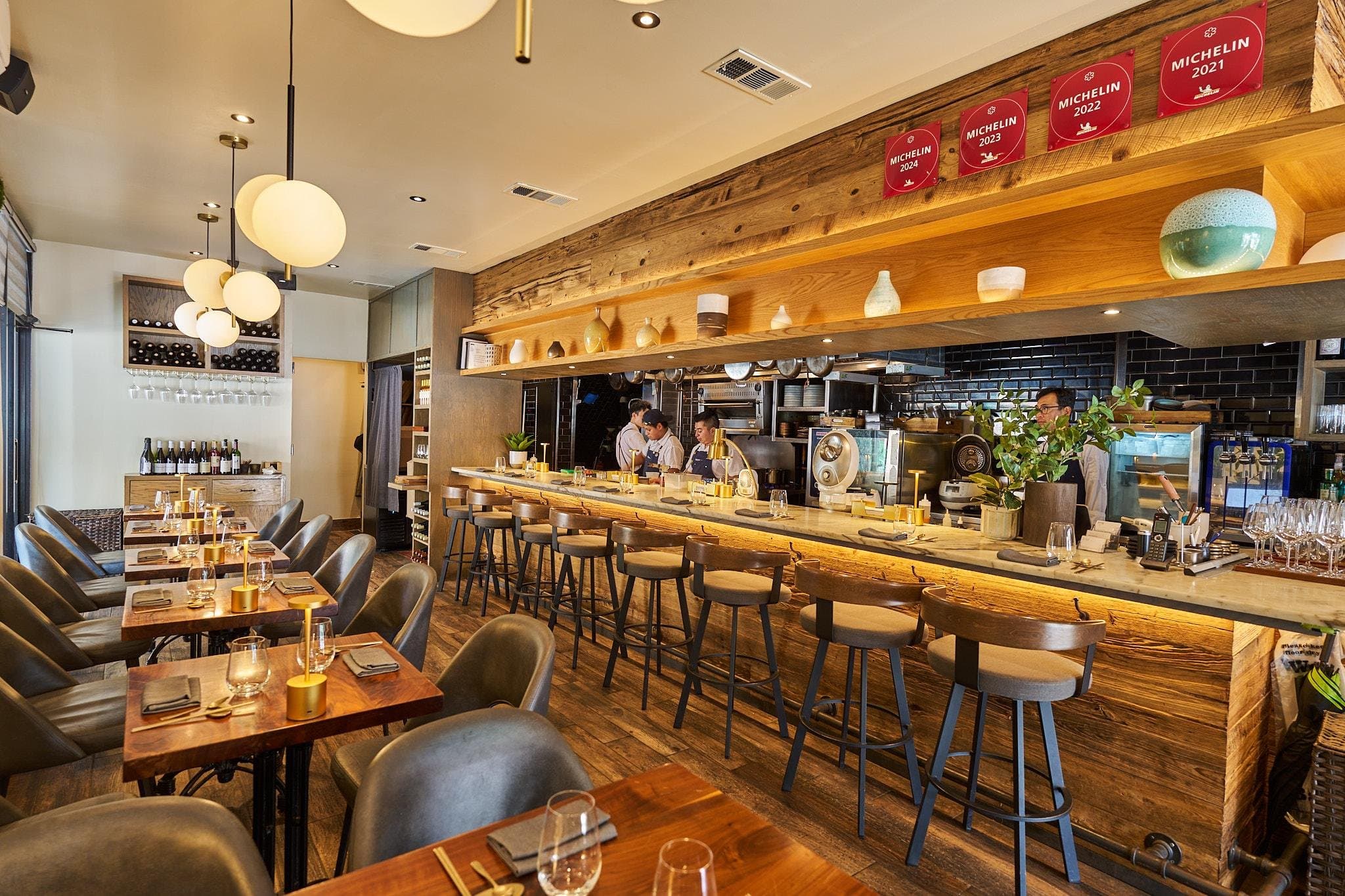 Kochi’s Hell’s Kitchen dining room with wood tables, chef’s counter, open kitchen, and Michelin Guide plaques on the wall