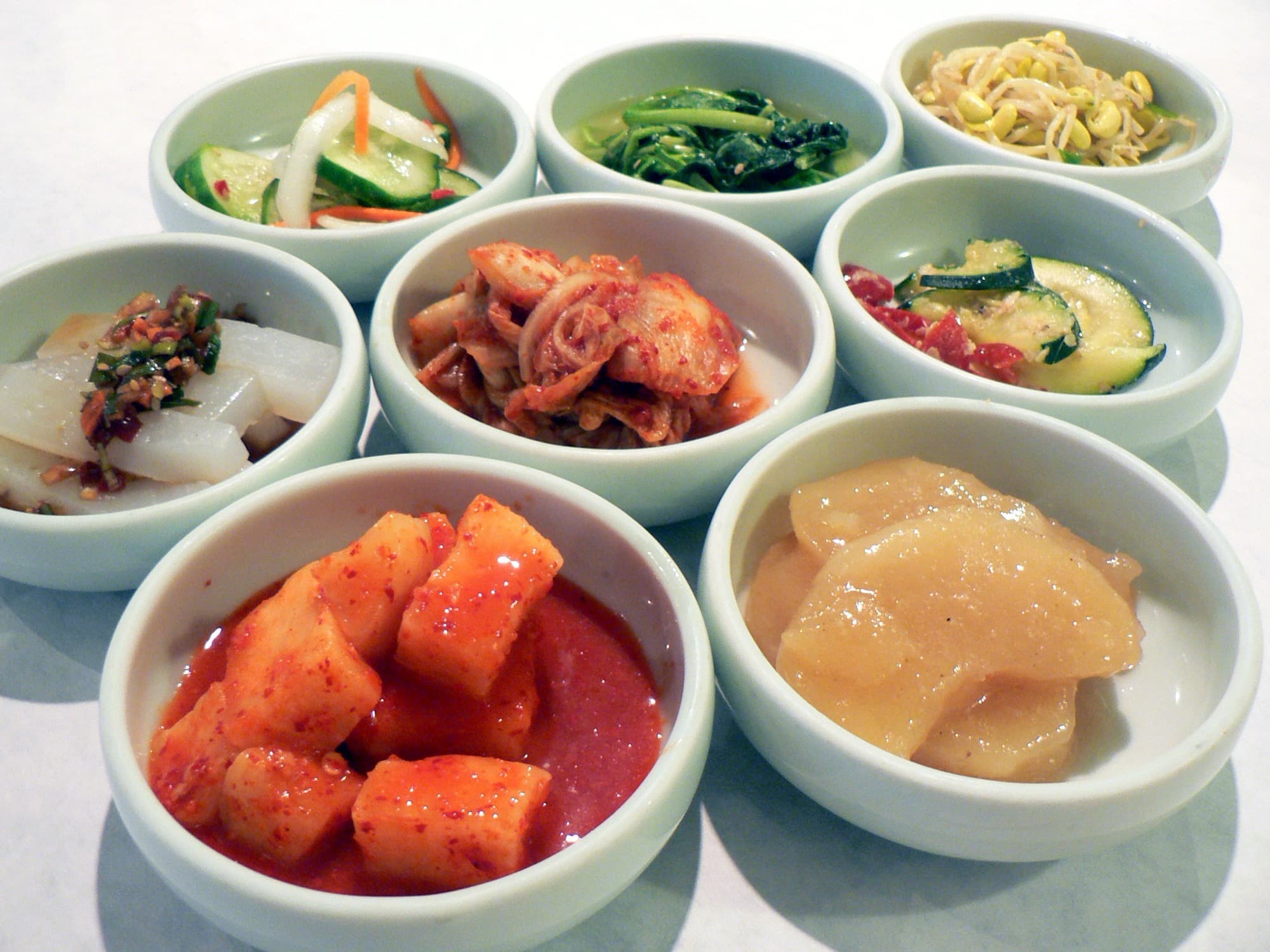 Eight small white bowls of Korean banchan arranged in rows on a white table