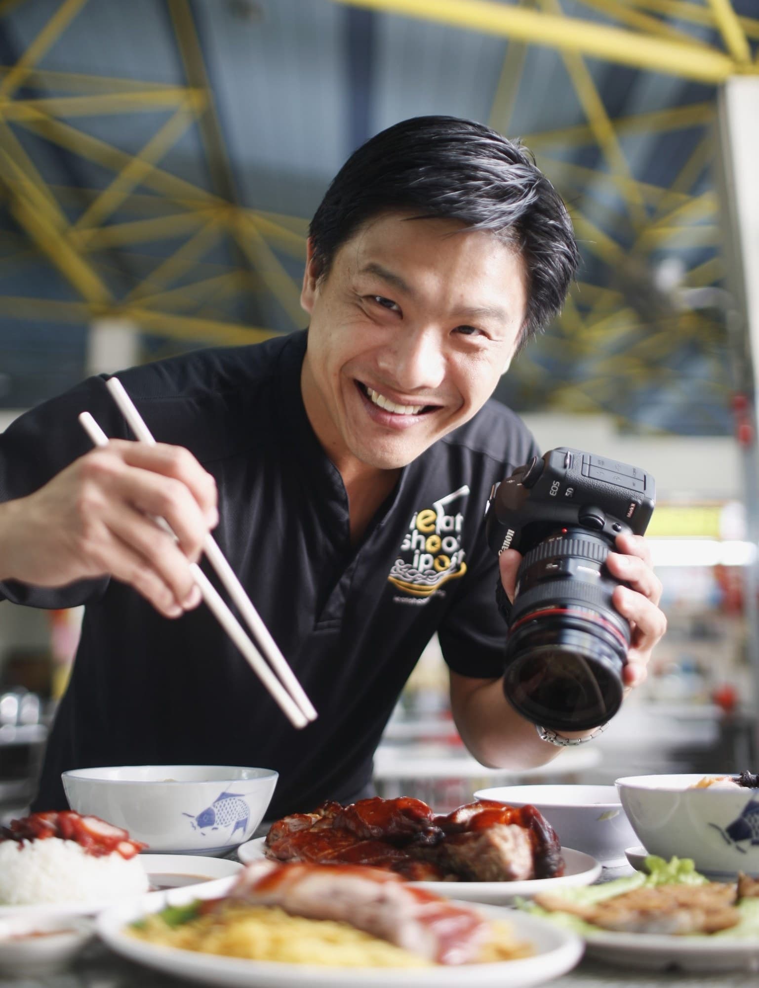 Leslie Tay smiling at a hawker table, holding chopsticks and a camera, with roast meat and rice in front of him