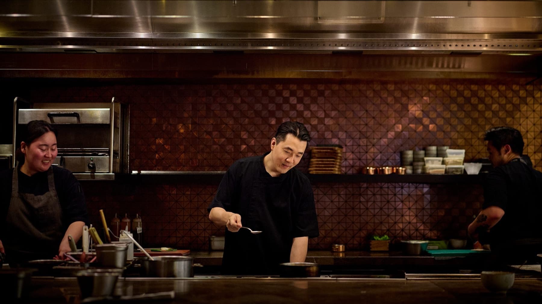 Chef Louis Han in a black chef’s shirt tasting from a spoon at a stainless-steel pass, with kitchen staff working beside him