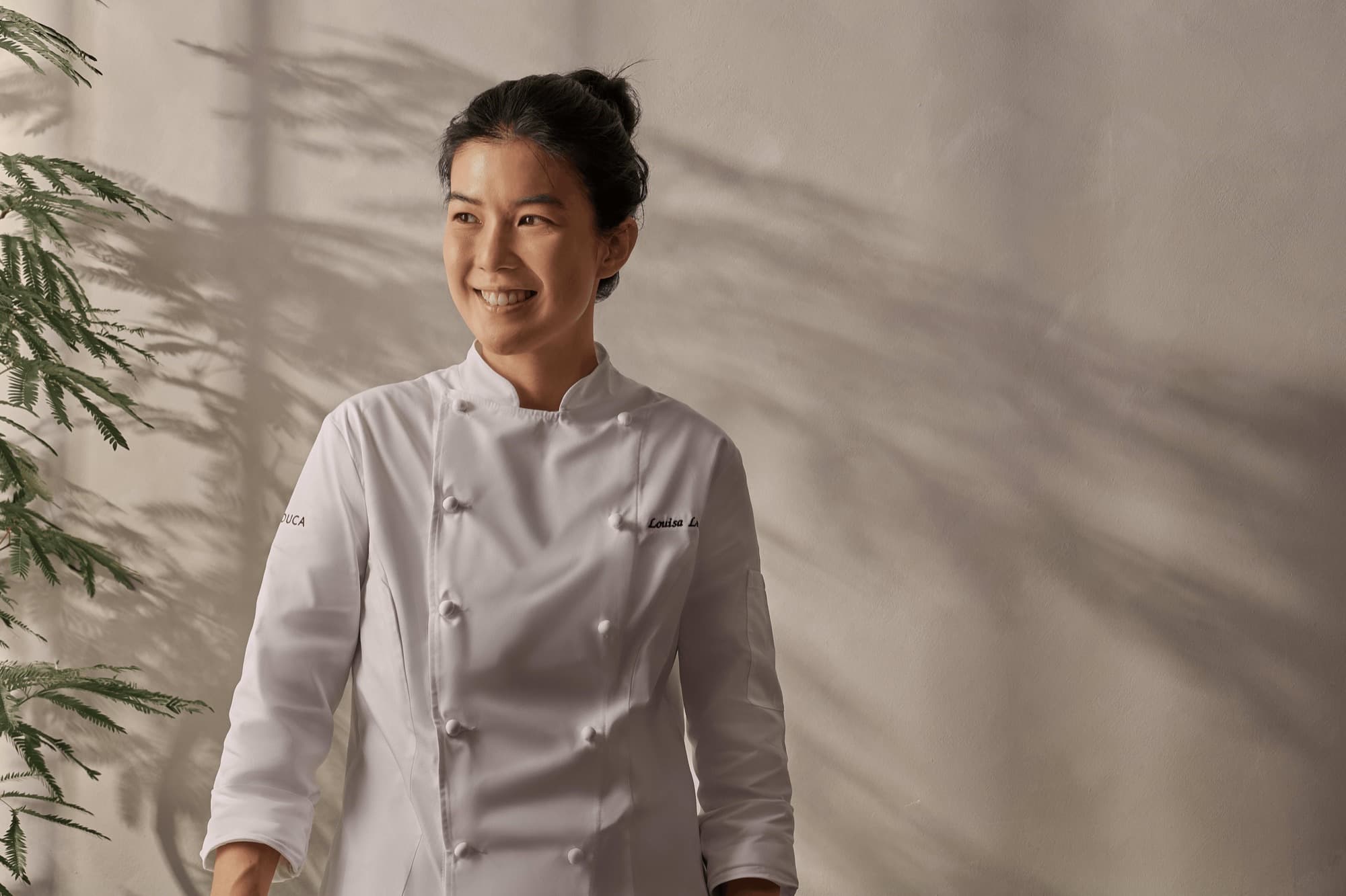 Pastry chef Louisa Lim in a white Loulouca chef’s coat, smiling against a sunlit wall
