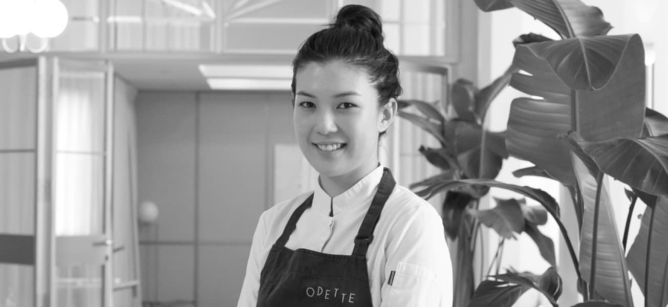 Louisa Lim smiling in a white chef’s coat and a dark Odette apron, standing in the restaurant dining room