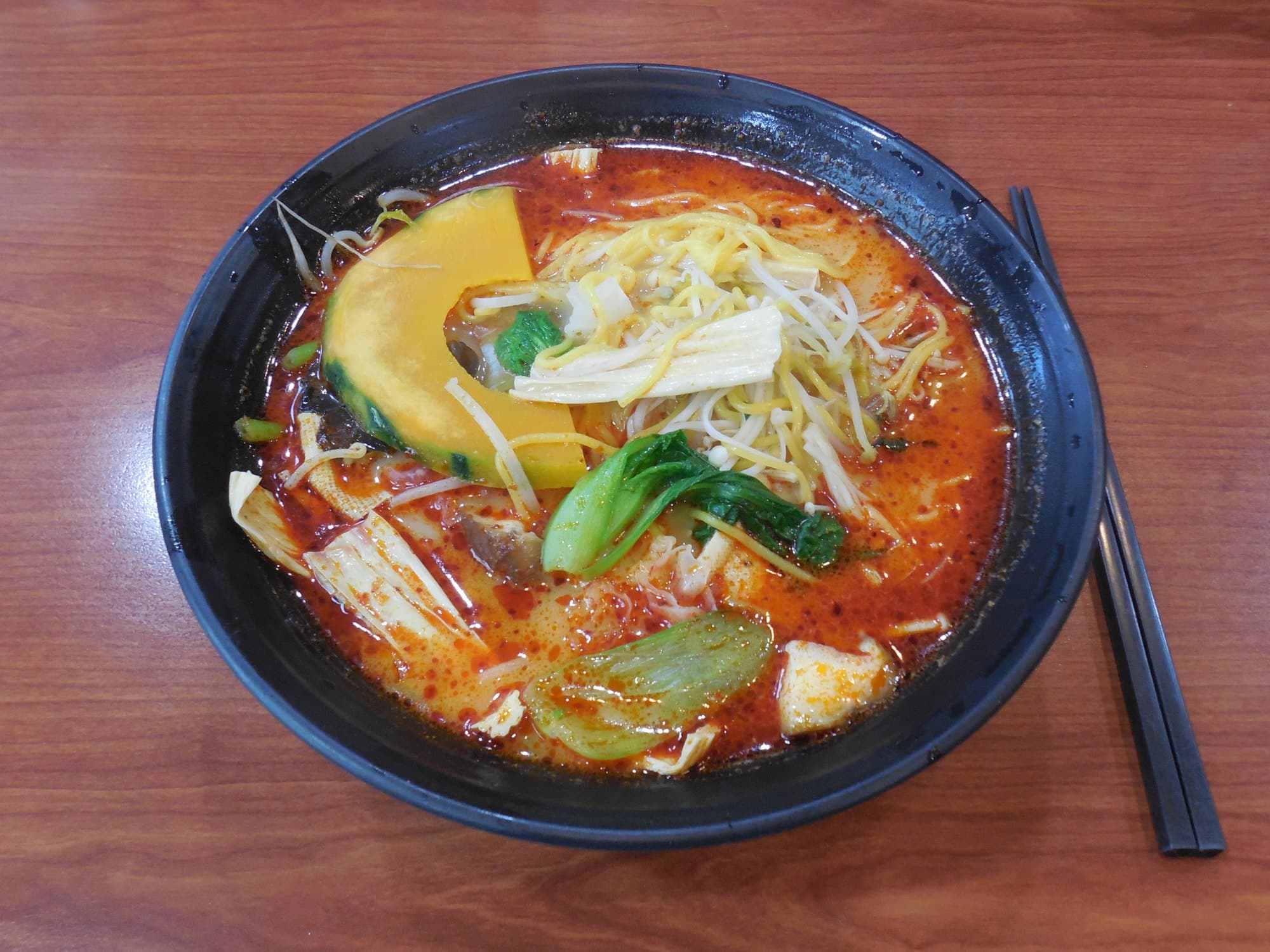 A black bowl of red malatang soup with kabocha squash, greens, bean sprouts, and noodles on a wood table in Seoul