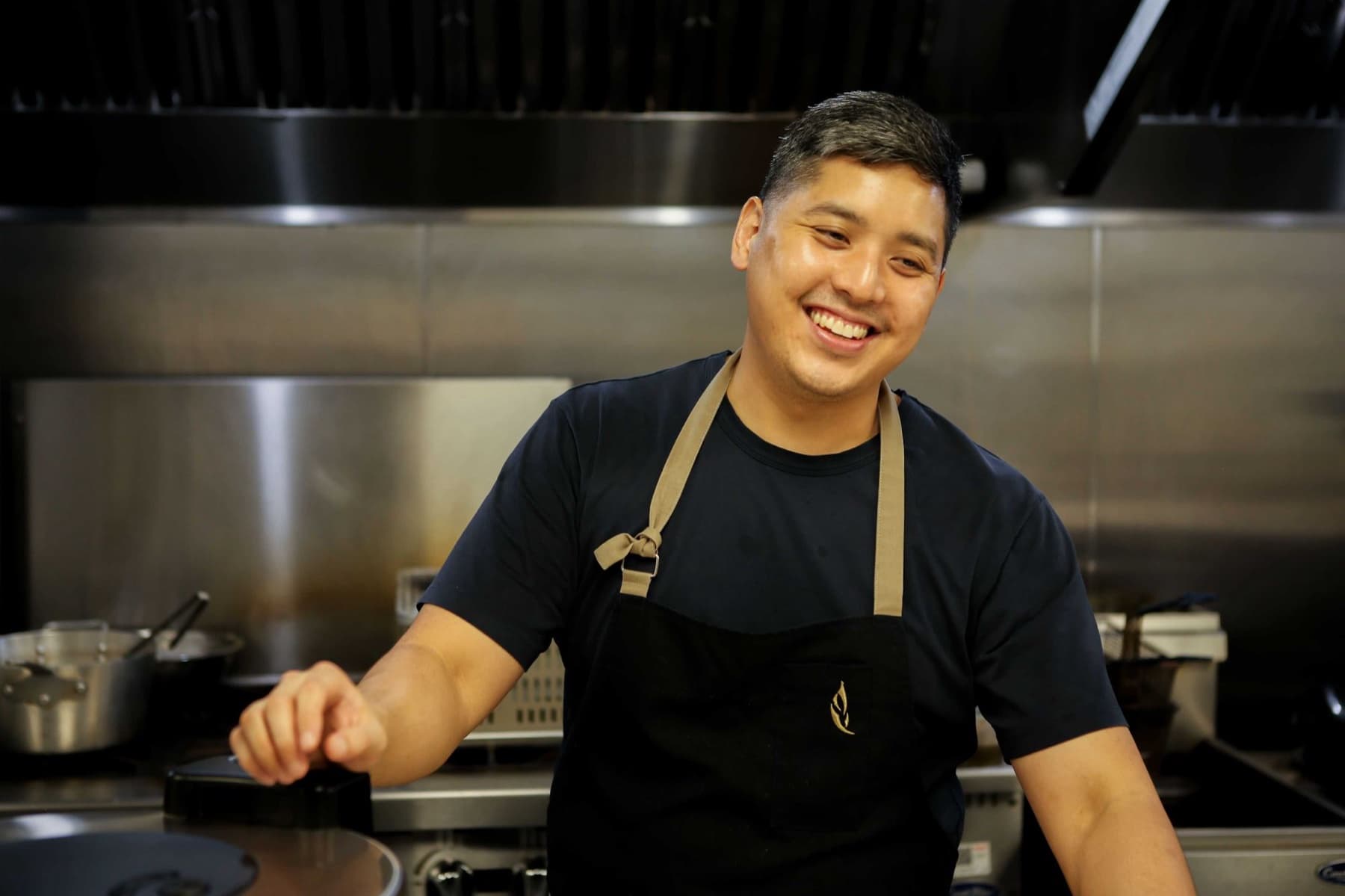Chef Kris Hoang in a black apron over a navy shirt, smiling in a commercial kitchen