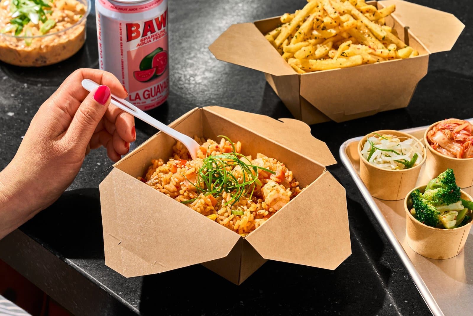 A hand with a plastic fork over a takeout box of kimchi fried rice with scallions, beside seasoned fries and three small cups of kimchi, bean sprouts, and broccoli