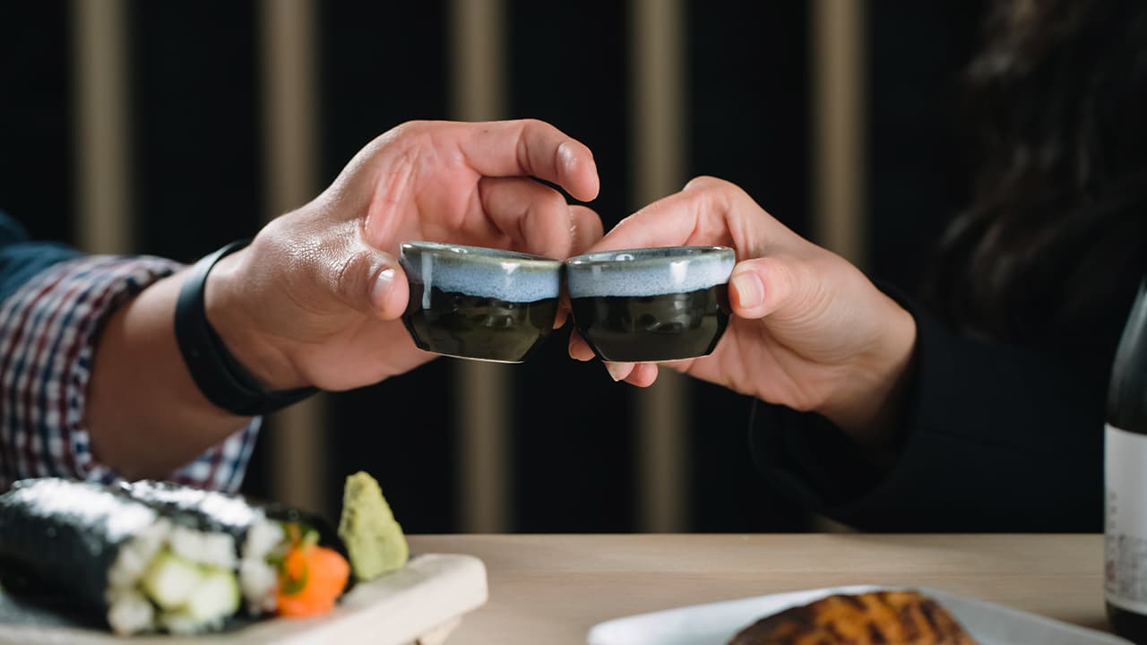 Two hands toasting small ceramic sake cups over a wooden board of temaki hand rolls