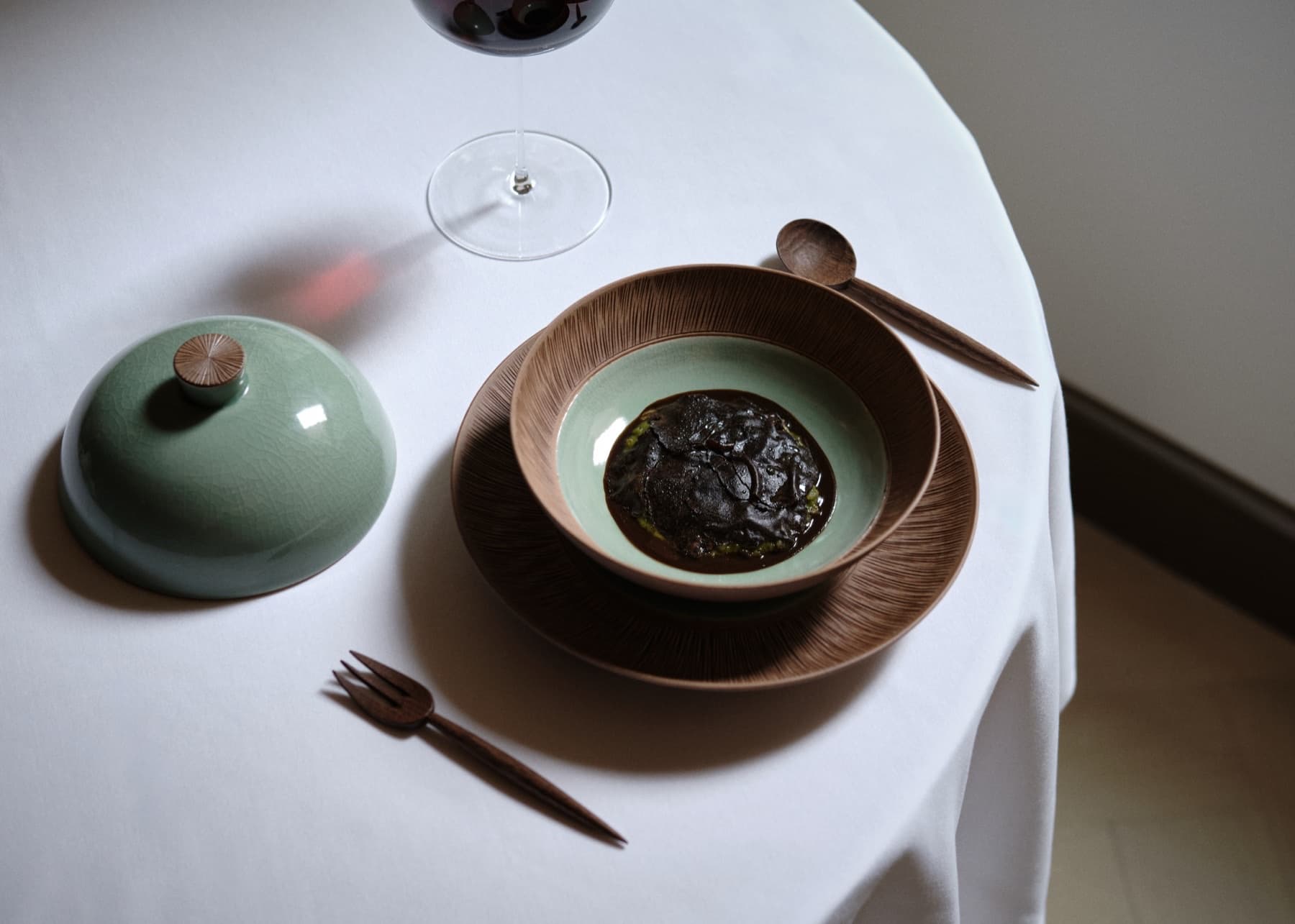 Oyatte’s spring green porridge under a blanket of soy-preserved black truffle, with wooden utensils and a celadon cloche on a white cloth