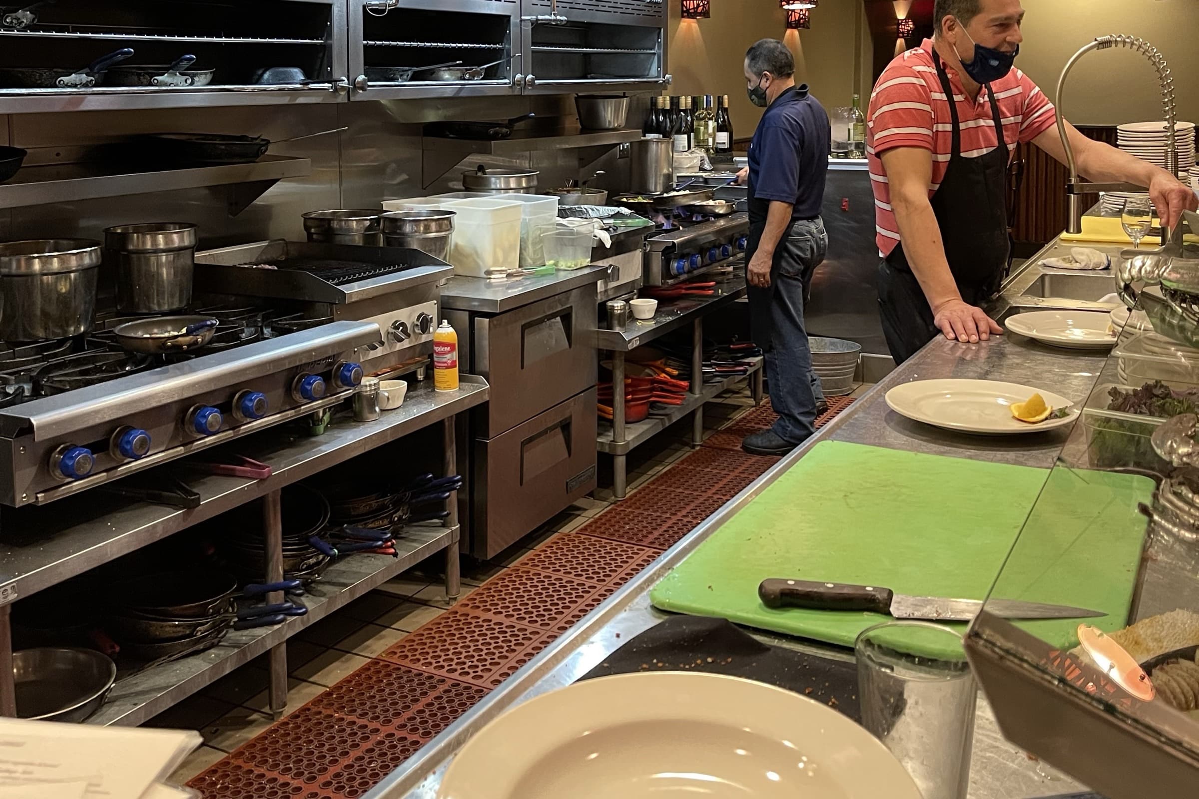 Restaurant kitchen prep counter with plates, cutting board, and line cooks at work