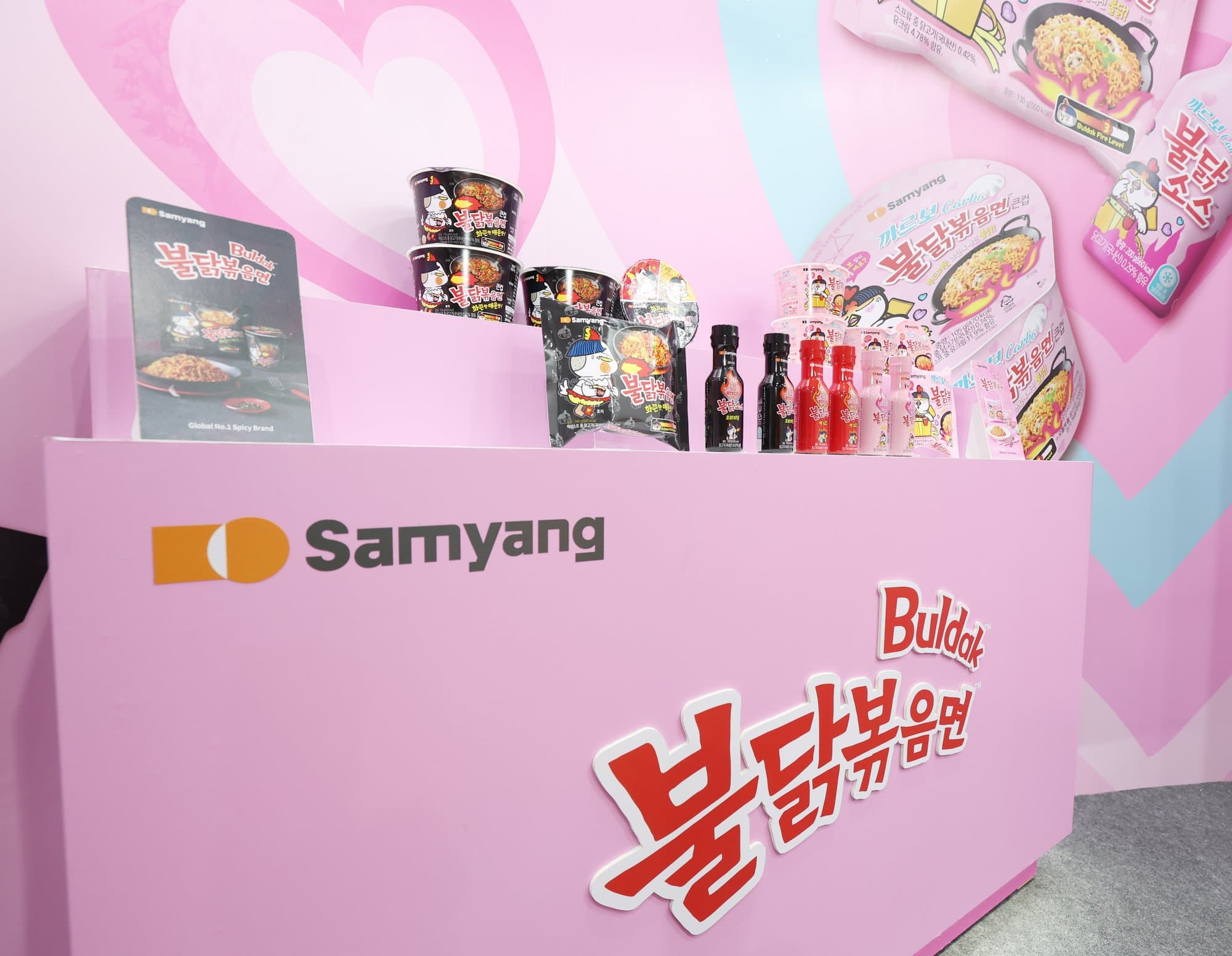 Pink Samyang booth at a ramen fair, with Buldak cups, sauce bottles, and the brand name on the counter