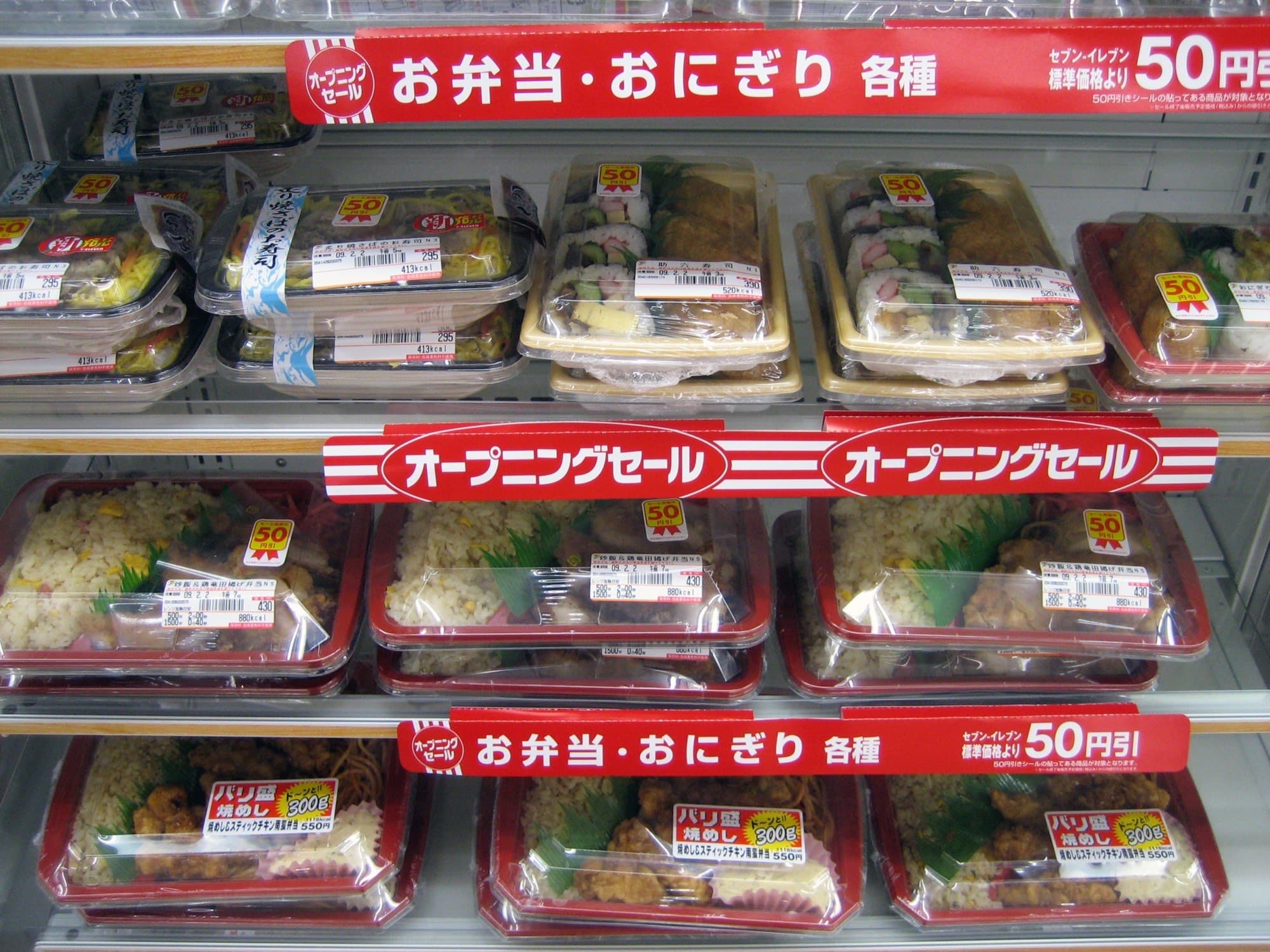 A Japanese 7-Eleven cold case stacked with sushi rolls and bento boxes behind red shelf banners