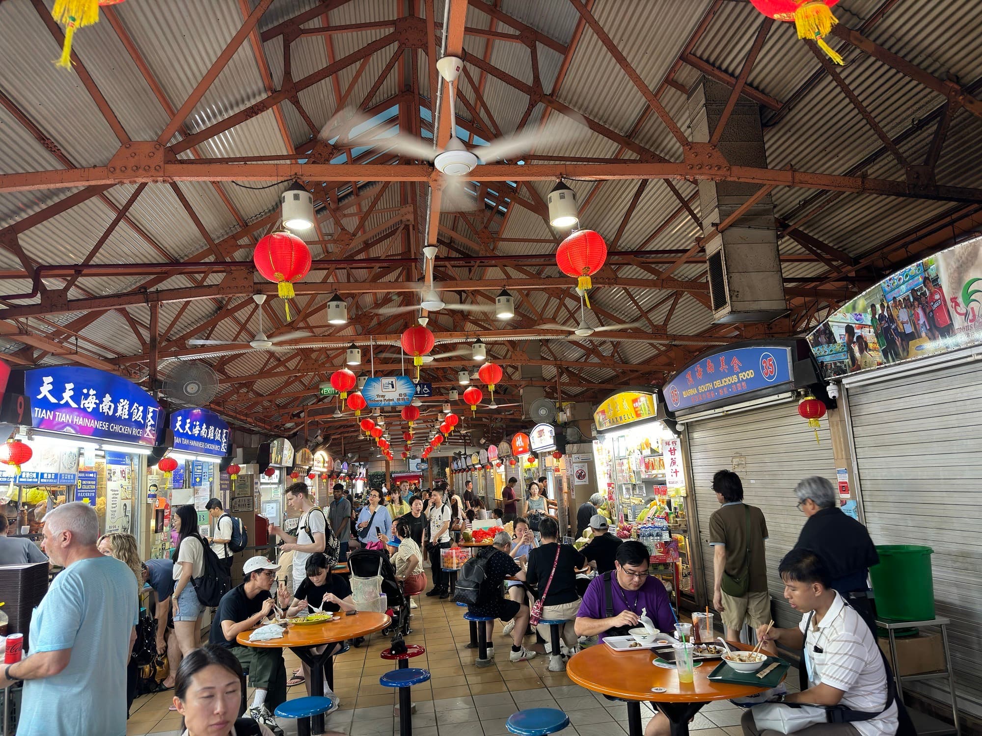 Diners at round tables inside Maxwell Food Centre under red lanterns, with Tian Tian Hainanese Chicken Rice signage along the stalls