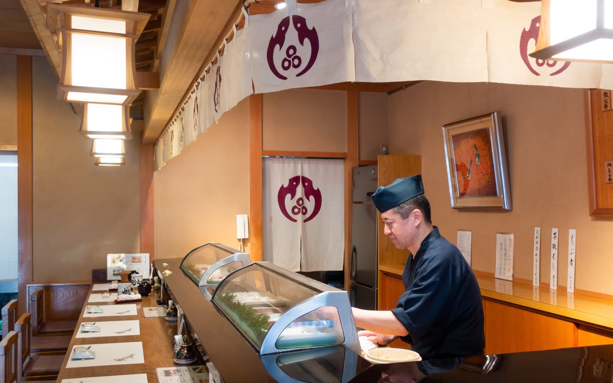 Sushi counter with empty place settings, fish case, and chef at work under warm lanterns