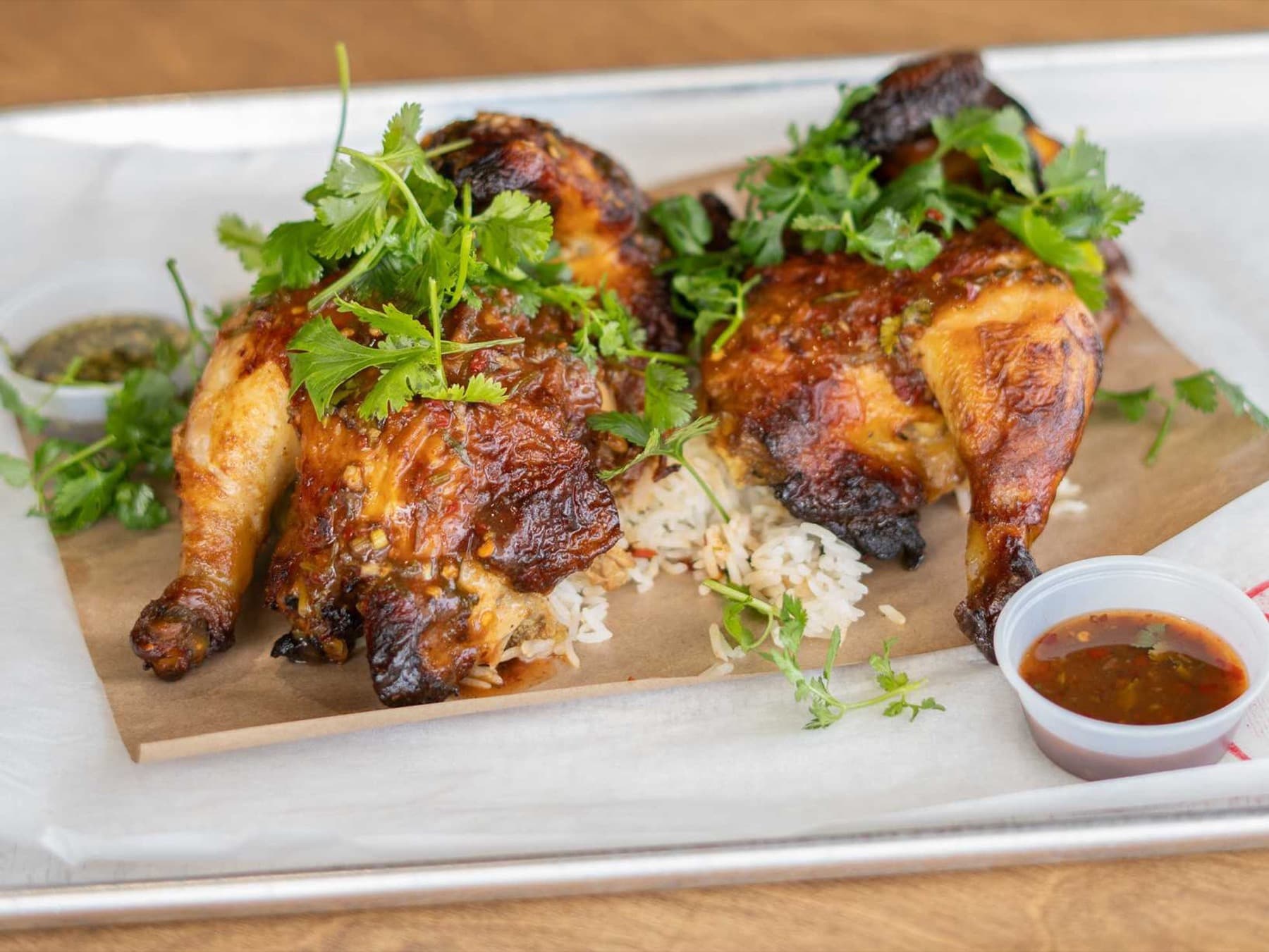 Halved Thai-style rotisserie chicken on jasmine rice with cilantro and two dipping sauces on a metal tray lined with paper