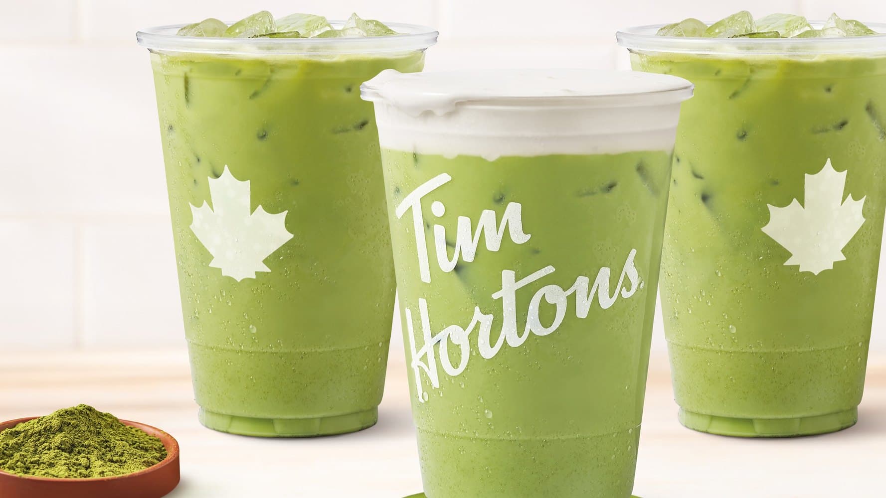 Three Tim Hortons iced matcha drinks on a light wood surface, one topped with cold foam, beside a small bowl of green matcha powder