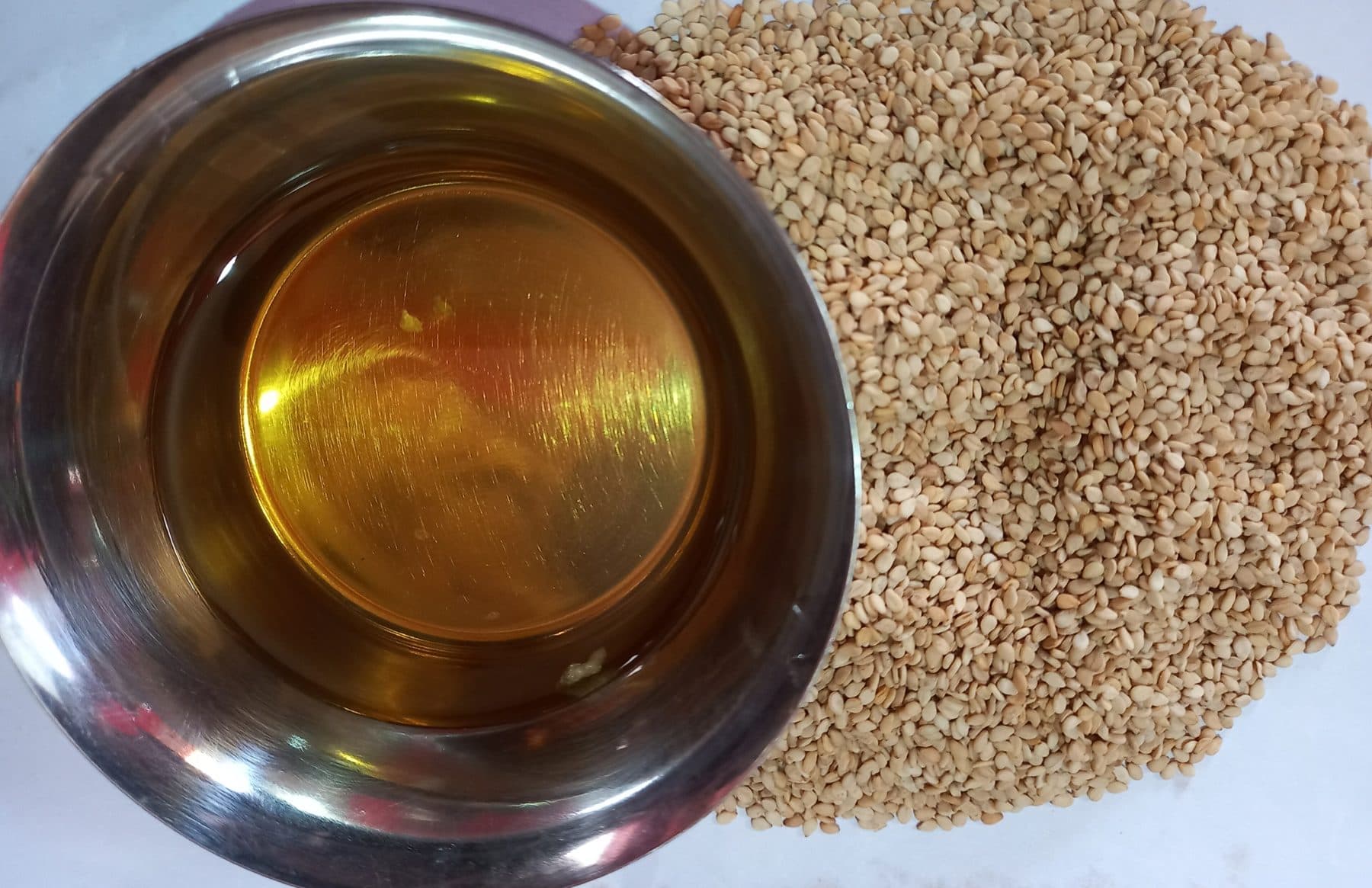A small stainless bowl of amber sesame oil next to a mound of toasted sesame seeds on a white surface