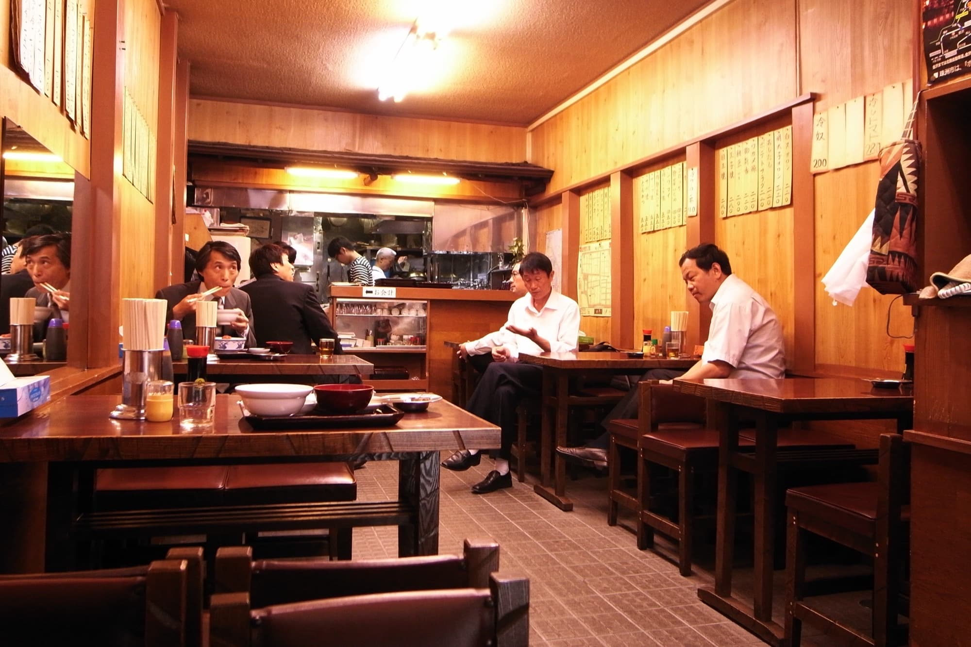 A narrow Tokyo izakaya with wooden walls, hanging menu strips, and diners at small tables
