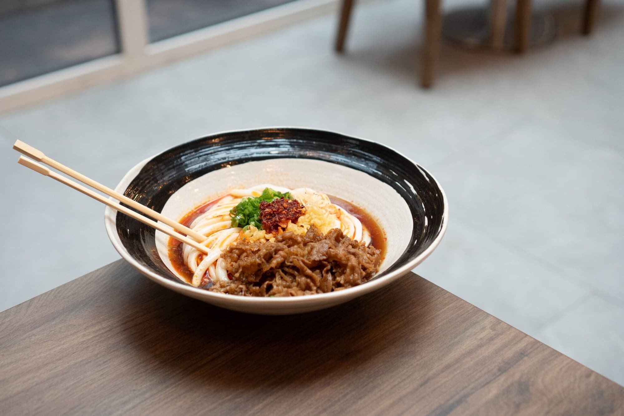 Spicy cold beef udon in a black-rimmed bowl, with chili oil, scallions, and tempura flakes, on a wood table